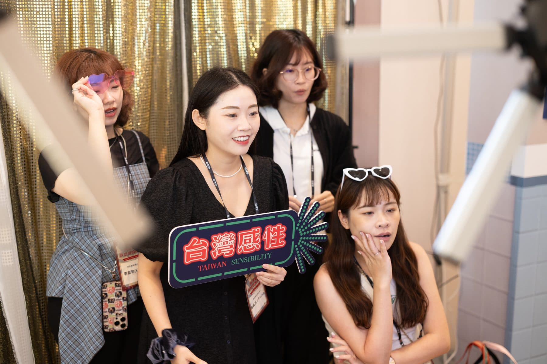Four guests posing in front of a gold sequin backdrop with neon-style sign props and heart glasses